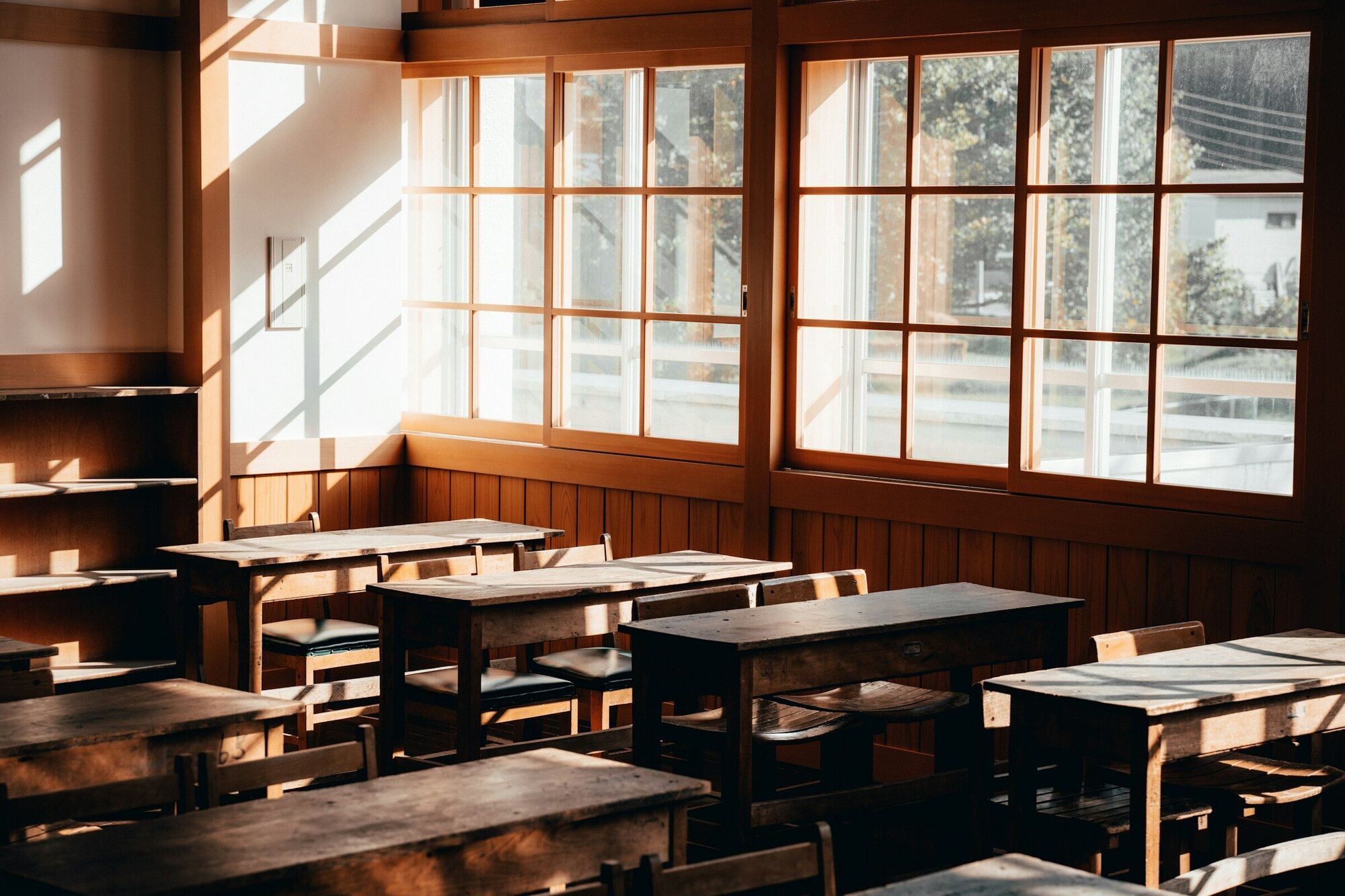 A classroom of wooden desks beneath tall windows, lit by afternoon sun.