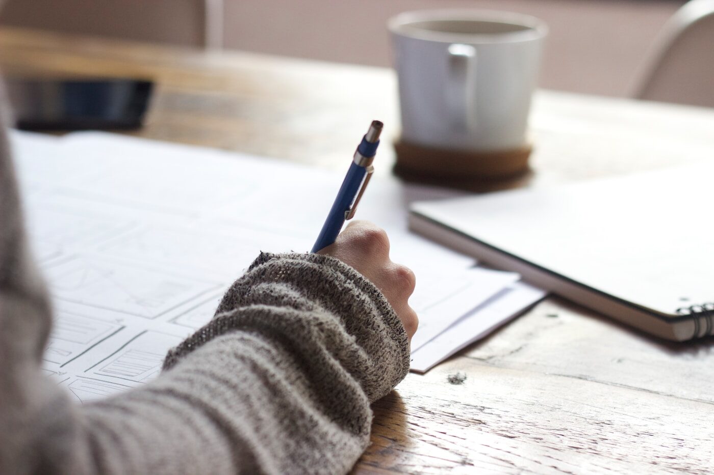 A hand filling in a printed form on a wooden desk beside a cup of coffee.